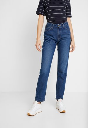 Personne portant un jean droit bleu foncé, un haut noir à manches courtes avec des rayures blanches, et des baskets blanches debout sur un sol blanc.