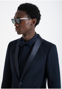 Tuxedo blu navy con rever in satin, papillon abbinato e taglio aderente; modello che indossa occhiali da sole scuri e angolari. Texture liscia, design su misura.