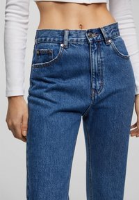 Los pantalones vaqueros de tiro alto en denim azul presentan costuras tonal, un cierre de cremallera y bolsillos frontales. La tela tiene un acabado ligeramente texturizado.