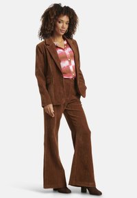 Brown corduroy suit with a fitted blazer and flared trousers. Underneath, a pink and orange patterned shirt. Brown heeled ankle boots.