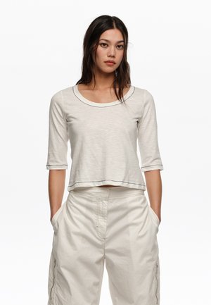 Joven con pelo largo y oscuro que lleva una camiseta beige clara de tres cuartos de manga y pantalones beige holgados, con las manos en los bolsillos, fondo neutro.