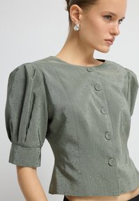 Eine Frau trägt eine salbeigrüne Kurzarmbluse mit puffartigen Schultern, mit stoffbedeckten Knöpfen und dezenten metallischen Sprenkeln.