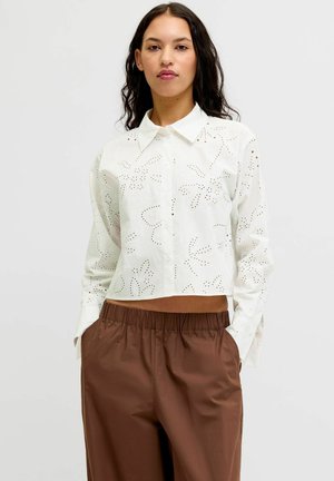 Kvinde med langt mørkt hår iført hvid øjlet cropped bluse og løse brune bukser, står med hænderne i lommerne mod en ensfarvet baggrund.