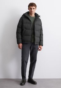 Giacca a vento nera con design trapuntato, cappuccio aderente e tasche laterali. Indossata sopra un maglione verde e pantaloni grigi su misura, abbinata a stivali neri.