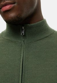 Grüner Strickpullover mit hohem Kragen und Reißverschluss. Der Stoff ist strukturiert mit gerippten Details und verfügt über einen metallischen Reißverschlussakzent.