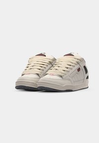 Beige athletic shoes with textured material, rounded toe, thick laces, and black accents on the sides and sole. Red logo detail on tongue.