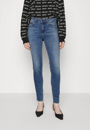 Nainen, joka pukeutunut keskisävyisiin skinny-farkkuihin, mustiin kärjestä teräviin slingback-kenkiin ja mustaan pitkähihaiseen toppiin, jossa on valkoinen "LIU JO"-tekstikuvio.