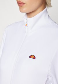 Valge tõmblukujope, millel on kõrge krae, tikandiga mitmevärviline logo ning soonilised detailid krae ja varrukaotstes. Sile tekstuur.