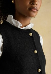 Gilet di lana nera con superficie texture, dotato di tre bottoni dorati. Indossato sopra una blusa bianca con ruches al colletto.