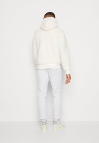 Man med vit fluffig huvtröja, ljusgrå joggingbyxor och vita Nike-sneakers, som står vänd mot en vit vägg.