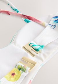 Bela tkanina z rožnim vzorcem z turkiznimi in roza trakovi, z zlato kovinsko opremo in gladko teksturo. Izpostavljeni detajli nastavljivih trakov.