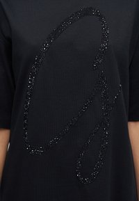 Μαύρο βαμβακερό t-shirt με εμφανές γυαλιστερό μαύρο σχέδιο με χάντρες σε σχήμα στυλιζαρισμένου γράμματος στην πρόσοψη.