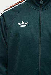 Veste zippée vert foncé avec des bandes rouges, blanches et noires sur l'épaule et un logo trefoil blanc d'Adidas sur la poitrine.
