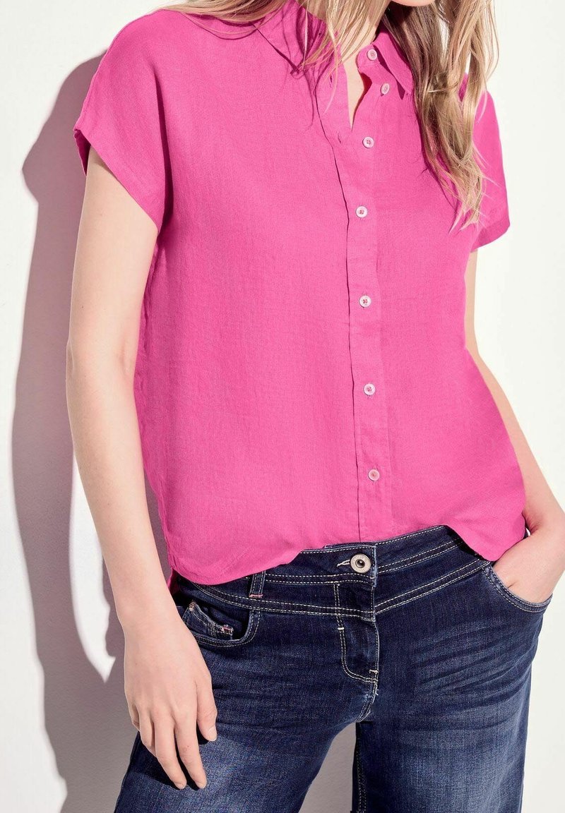 Camicia fucsia a maniche corte con colletto. Realizzata in tessuto leggero, presenta una patta frontale e un orlo curvo. Abbinata a jeans scuri.