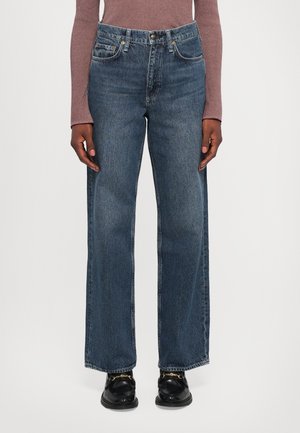 Mannequin portant un jean bleu taille haute à jambe droite avec poches avant, associé à un haut mauve à manches longues et des mocassins noirs avec un détail chaîne dorée.