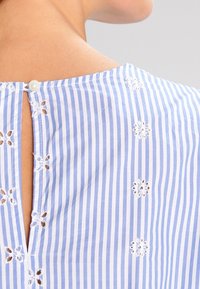 Blusa a righe blu e bianche con chiusura a bottoni, schiena scoperta e dettagli in ricamo floreale sul tessuto. Texture liscia.