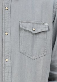 Cămașă din denim gri deschis, cu închidere prin butoane, având un buzunar pe piept și butoane metalice argintii pentru închidere. Textură moale și croială standard.