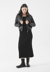 Žena v černé kožené bundě přes černé ribbed midi šaty a černých kotníkových botách, stojící na bílém pozadí.