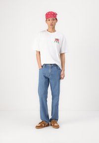 Tricou alb cu mânecă scurtă și un grafic colorat, blugi albaștri drepți, adidași maro și o bandană roșie pe frunte.