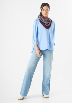 Chemise bleu clair à manches retroussées, associée à un jean large délavé et à une écharpe à motifs gris et bordeaux. Baskets blanches.
