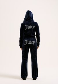 Μπλέ βελούδινο κοστούμι φόρμας με κουκούλα, διακοσμημένο με "Juicy" από ρουμπίνια στην πλάτη του σακακιού και των παντελονιών. Χαλαρή εφαρμογή με μακριά μανίκια.