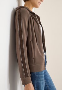 Brązowa bluza z kapturem na suwak, z podszytym kapturem, ozdobiona fakturowanym wzorem i napisem "FOCUS AHEAD" w kolorze brązowym wzdłuż bocznych szwów.