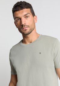 Lichtgrijze T-shirt met korte mouwen en een ronde hals, voorzien van een subtiele zwarte gestreepte logo op de borst. Zachte textuur, slimfit ontwerp.