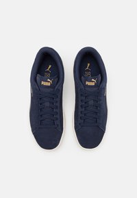 Marina mockasiner i mocka med platta snören, vadderad krage, guldlogotypdetaljer och strukturerad gummisula. Klassisk låg topp-design.