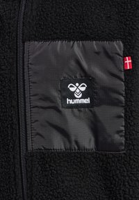 Veste en polaire noire avec une poche poitrine en nylon noir lisse arborant un logo blanc. Détail avec le drapeau danois rouge sur le côté. Fermeture à glissière.
