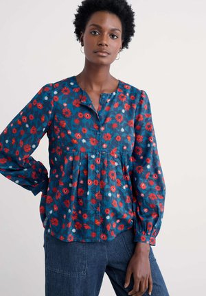 Bloes met bloemenprint in donkerblauw, met rode en witte bloemen, plooien en een ronde halslijn met knoopsluiting. Lange pofmouwen.