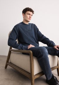 Granatowy sweter z teksturowanymi dzianinowymi rękawami i czarnym logo. Noszony z ciemnymi dżinsami, siedząc na krześle z drewnianymi akcentami w minimalistycznym otoczeniu.