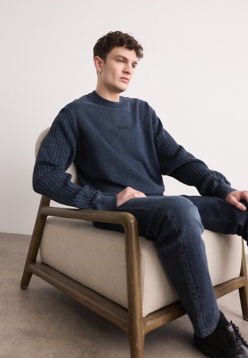 Granatowy sweter z teksturowanymi dzianinowymi rękawami i czarnym logo. Noszony z ciemnymi dżinsami, siedząc na krześle z drewnianymi akcentami w minimalistycznym otoczeniu.