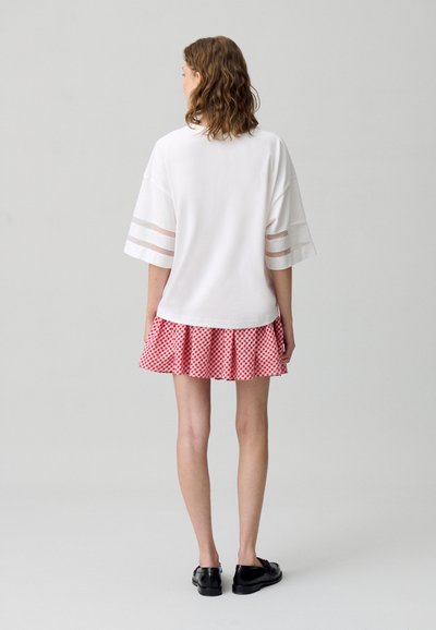 T-shirt bianco oversize con dettagli in strisce trasparenti, abbinato a una gonna a campana a quadretti rossi. Scarpe nere. Design semplice e casual.