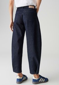 Pantalon large en denim bleu foncé à taille haute, avec poches arrière et étiquette en cuir à la taille. Texture lisse, coupe droite.