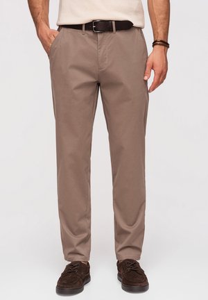 Homme portant un pantalon chino beige, une ceinture marron foncé et des chaussures en daim marron foncé à lacets, debout, bras le long du corps, sur un fond uni.