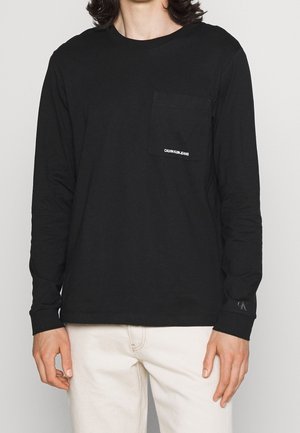Personne portant une chemise noire à manches longues avec une poche poitrine étiquetée "Calvin Klein Jeans" et un pantalon beige sur un fond uni.