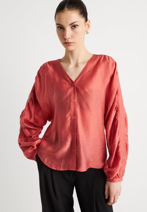 Koralsatinbluse med V-hals, lange puffede ærmer og knaplukning. Glat tekstur med subtil glans, kombineret med sorte bukser.