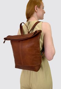 Brązowy skórzany plecak z zamkiem na górze, gładka tekstura, regulowane paski i zamek błyskawiczny. Dyskretny logo marki z boku.