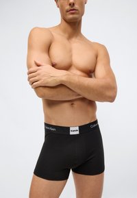 Muž bez trička se založenýma rukama, oblečený v černých boxerách Calvin Klein, stojící na neutrálním světlém pozadí.