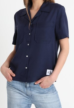 Femme portant une chemise à manches courtes bleu marine à boutons avec deux poches sur la poitrine et un jean bleu clair, les mains dans les poches.