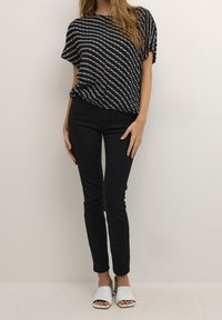 Femme portant un chemisier à manches courtes à motifs noirs et blancs, un pantalon noir slim et des sandales blanches à talons ouvertes, debout devant un fond uni.