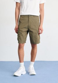 Bermudas de cargo en color caqui con dos bolsillos laterales, hechas de tela de algodón. Combinadas con una camiseta blanca y zapatillas blancas, de pie sobre una superficie azul.