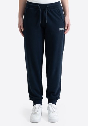 Persona che indossa pantaloni della tuta Lonsdale London blu navy con coulisse in vita e scarpe da ginnastica bianche, in piedi su uno sfondo semplice.