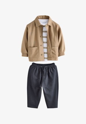 Beige Jacke mit Knopfleiste und zwei Vordertaschen über einem weiß-braun gestreiften Hemd, kombiniert mit dunkelgrauen Hosen mit elastischem Bund.