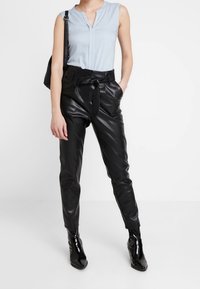 Donna con una blusa senza maniche di colore blu chiaro, pantaloni in pelle nera a vita alta con cintura, stivali neri alla caviglia e che porta una borsa a tracolla nera.