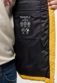 Zwarte en gele jasinterieur met een merklabel en gebruiksaanwijzingen. Materiaal is glad en heeft een winddichte en waterafstotende ontwerpeigenschap.
