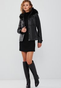 Giubbotto di pelle nera con cappuccio in pelliccia artificiale, due tasche frontali e chiusura con zip. Il modello indossa un vestito in maglia nero e stivali neri al ginocchio.