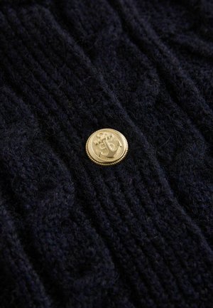 Marineblauer Strickpullover mit strukturierter Zopfmuster-Struktur und einem markanten goldfarbenen Ankerknopf mit geriffeltem Rand.