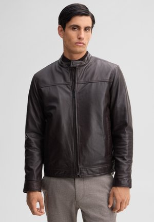 Mann trägt eine dunkelbraune Lederjacke mit Reißverschluss und Stehkragen sowie graue Hose und steht vor einem einfarbigen Hintergrund.
