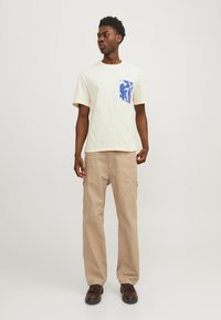 Creme kortærmet t-shirt med blå mønstret lomme, parret med beige cargo bukser og mørke sko. Modellen står let vendt til siden.
